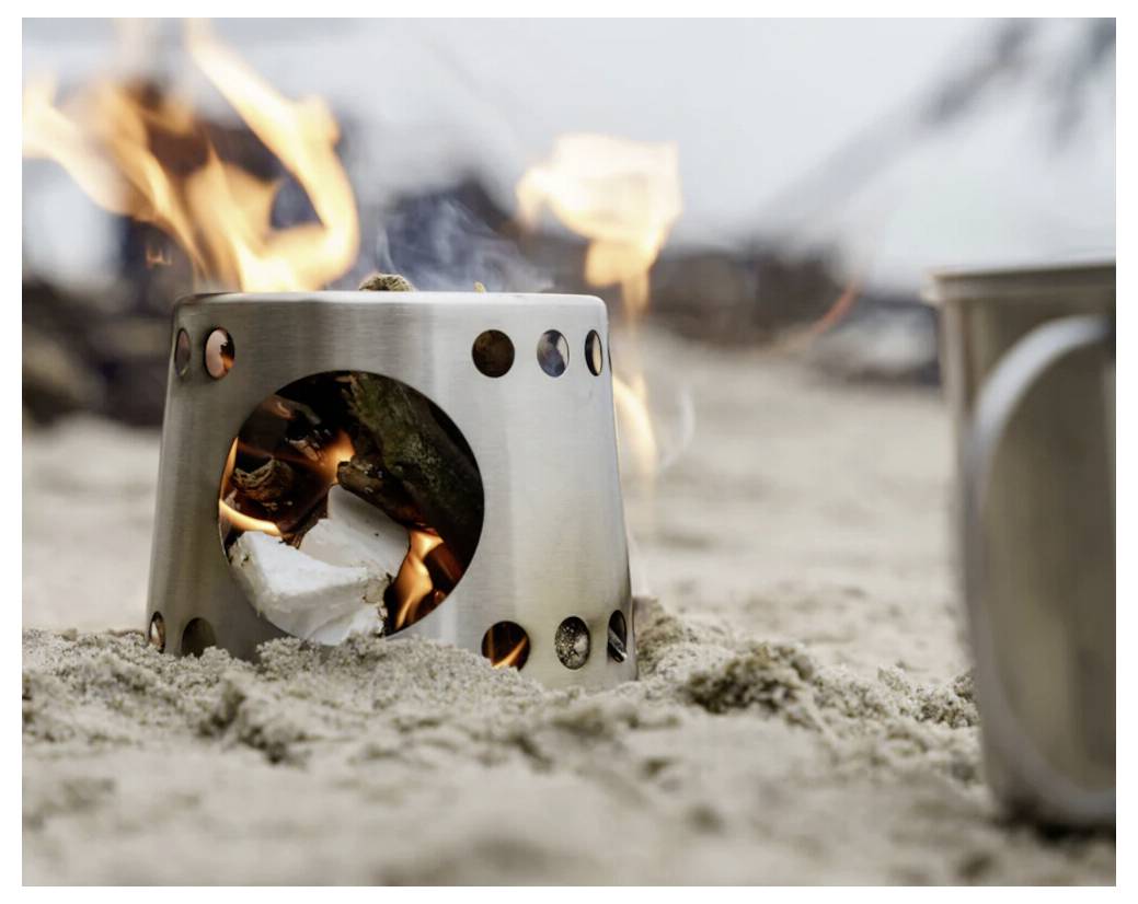 Kleiner Metallcampingkocher, der auf sandigem Boden brennt, mit sichtbaren Flammen durch die vordere Öffnung und Lüftungslöcher.