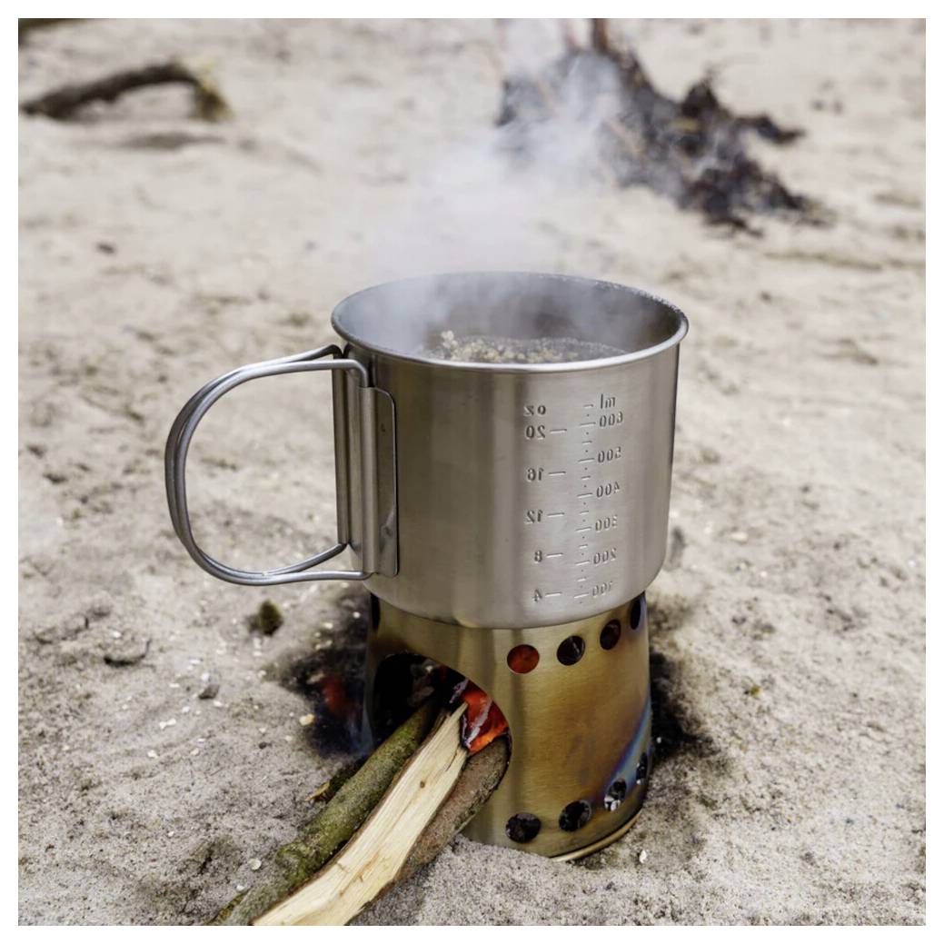 Edelstahlcampingtasse auf einem Holzfeuer-Rucksackwanderkochfeld im Sand, mit aufsteigendum Rauch und einem Stock, der die Flamme anfacht.