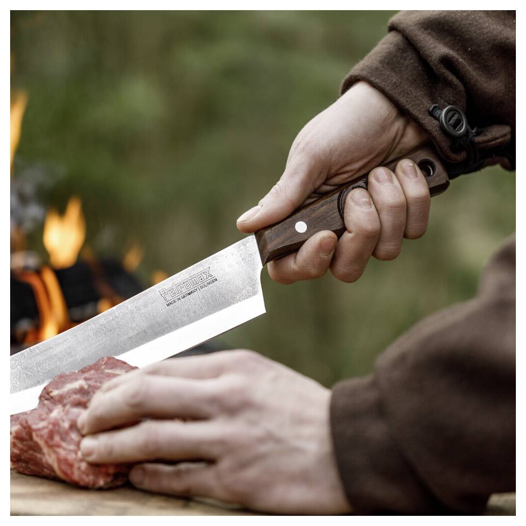 Eine Person, die ein großes Messer hält, schneidet rohes Fleisch neben einem Lagerfeuer im Freien.