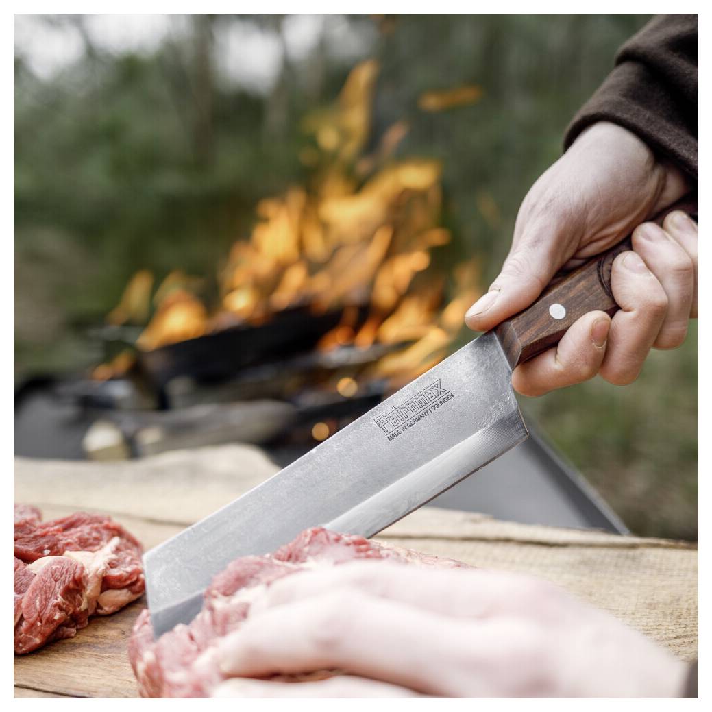 Eine Hand hält ein Hackemesser über rohes Fleisch auf einem Schneidebrett in der Nähe eines Lagerfeuers und bereitet Essen im Freien zu.