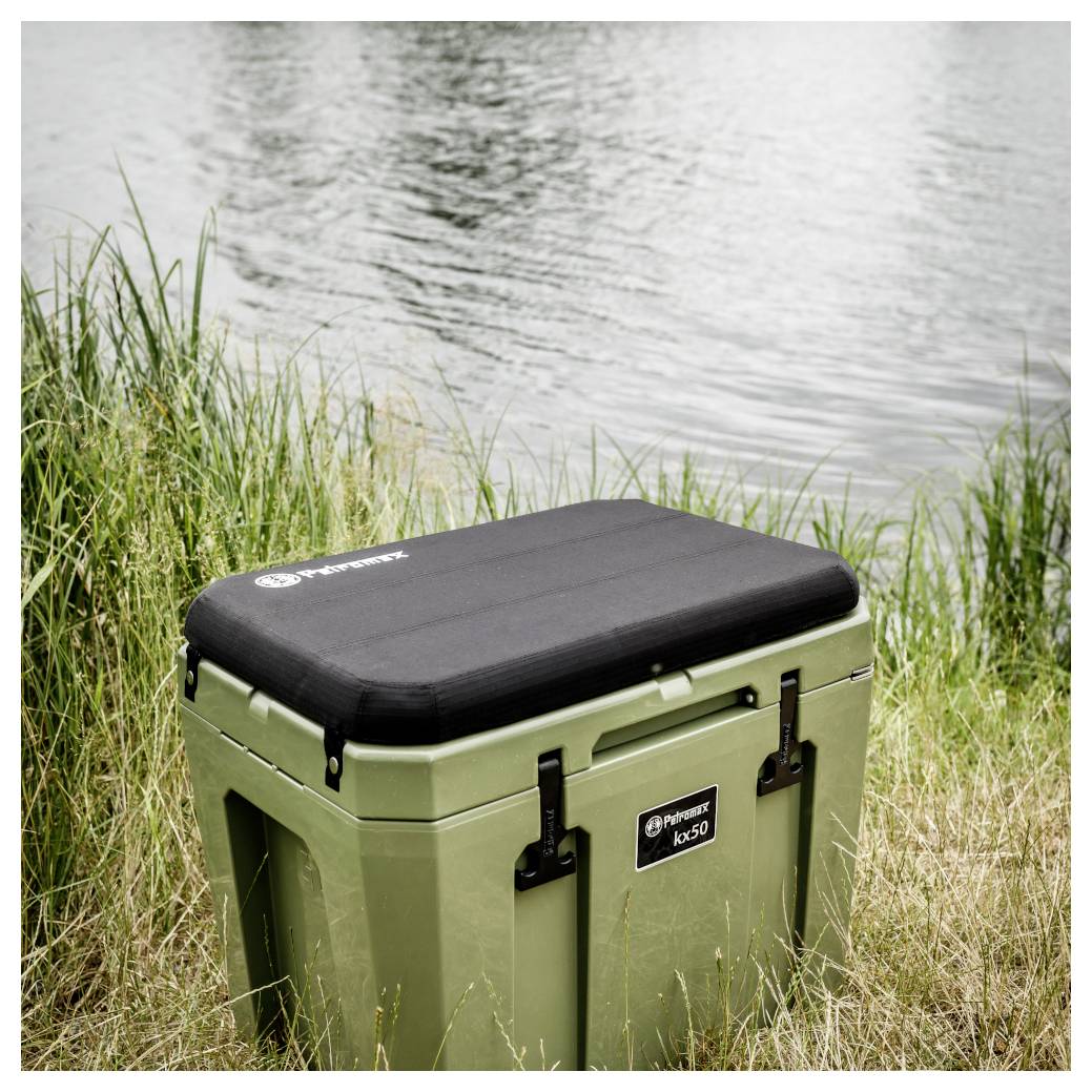 Ein olivgrüner tragbarer Kühlbox mit schwarzem Deckel sitzt im hohen Gras neben einem See.