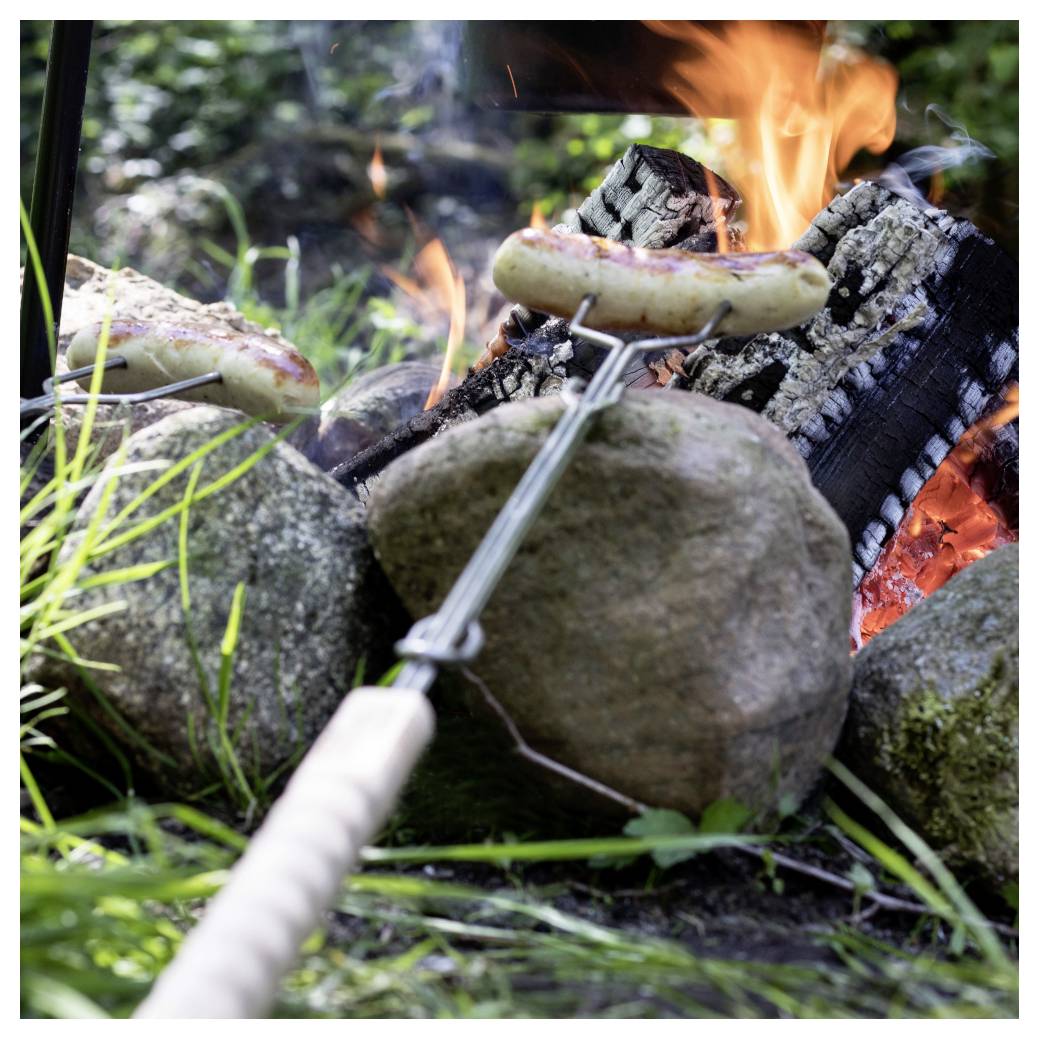 Würstchen braten auf Spießen über einem offenen Lagerfeuer zwischen Steinen und Baumstämmen in einer Waldumgebung.