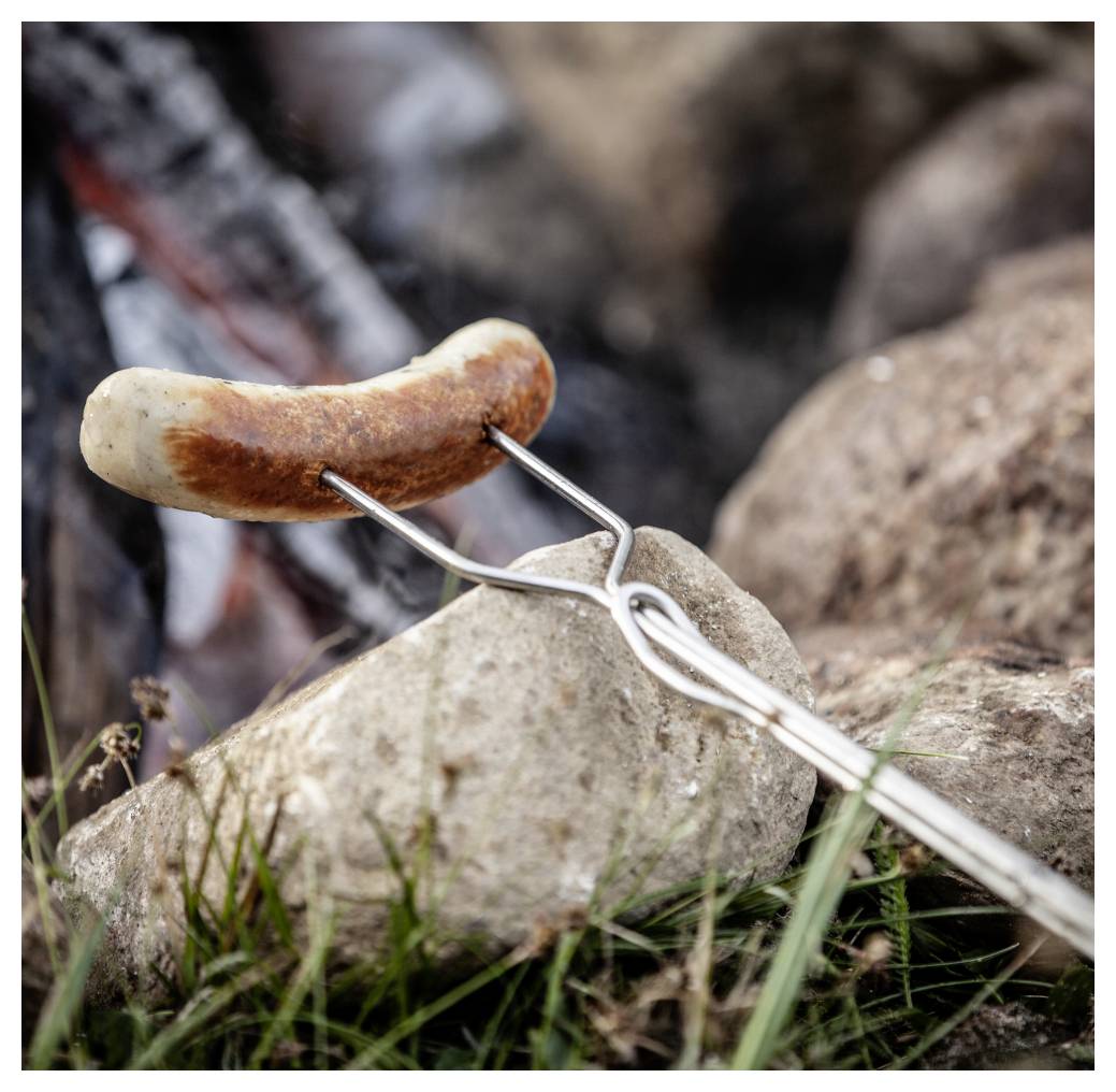 Eine Wurst wird auf einer Metallgabel über einem offenen Feuer geröstet, mit Steinen und Gras im Vordergrund.