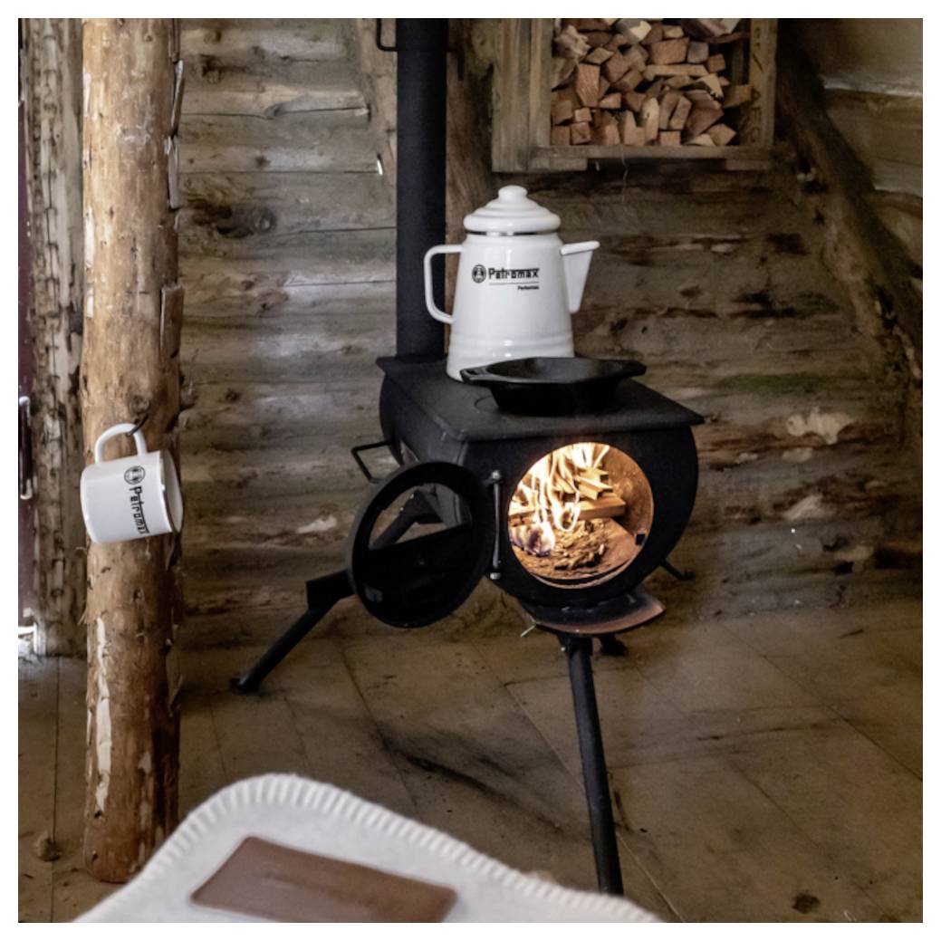 Holzofen, der eine rustikale Blockhütte heizt, mit einem Kessel oben drauf, einer Tasse auf einem Pfosten und Feuer, das durch die Ofentür glüht.