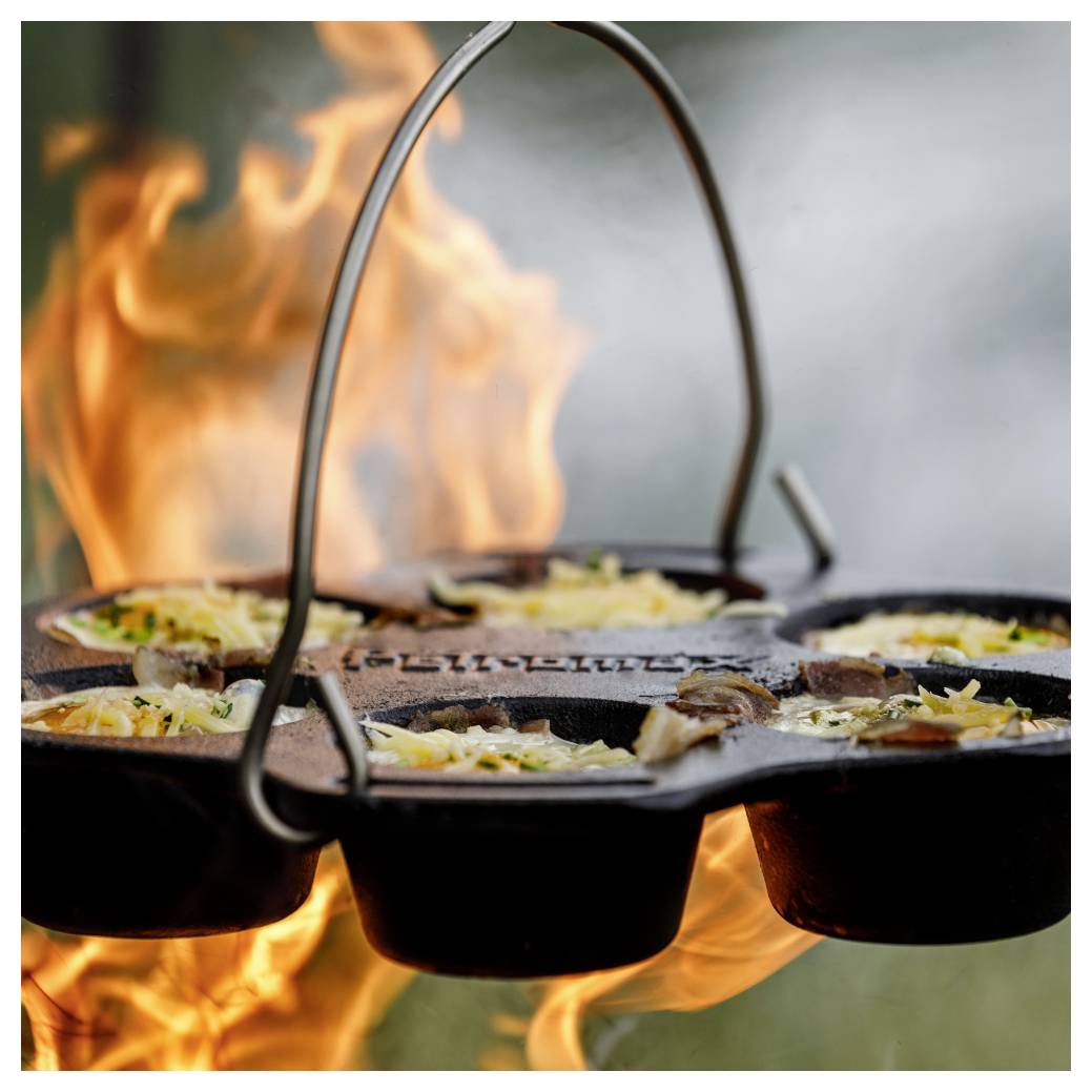 Gusseiserne Pfanne über offenem Feuer, in der mehrere gefüllte Eierschalen oder Omeletts zubereitet werden, mit Flammen, die um die Pfanne auflodern.