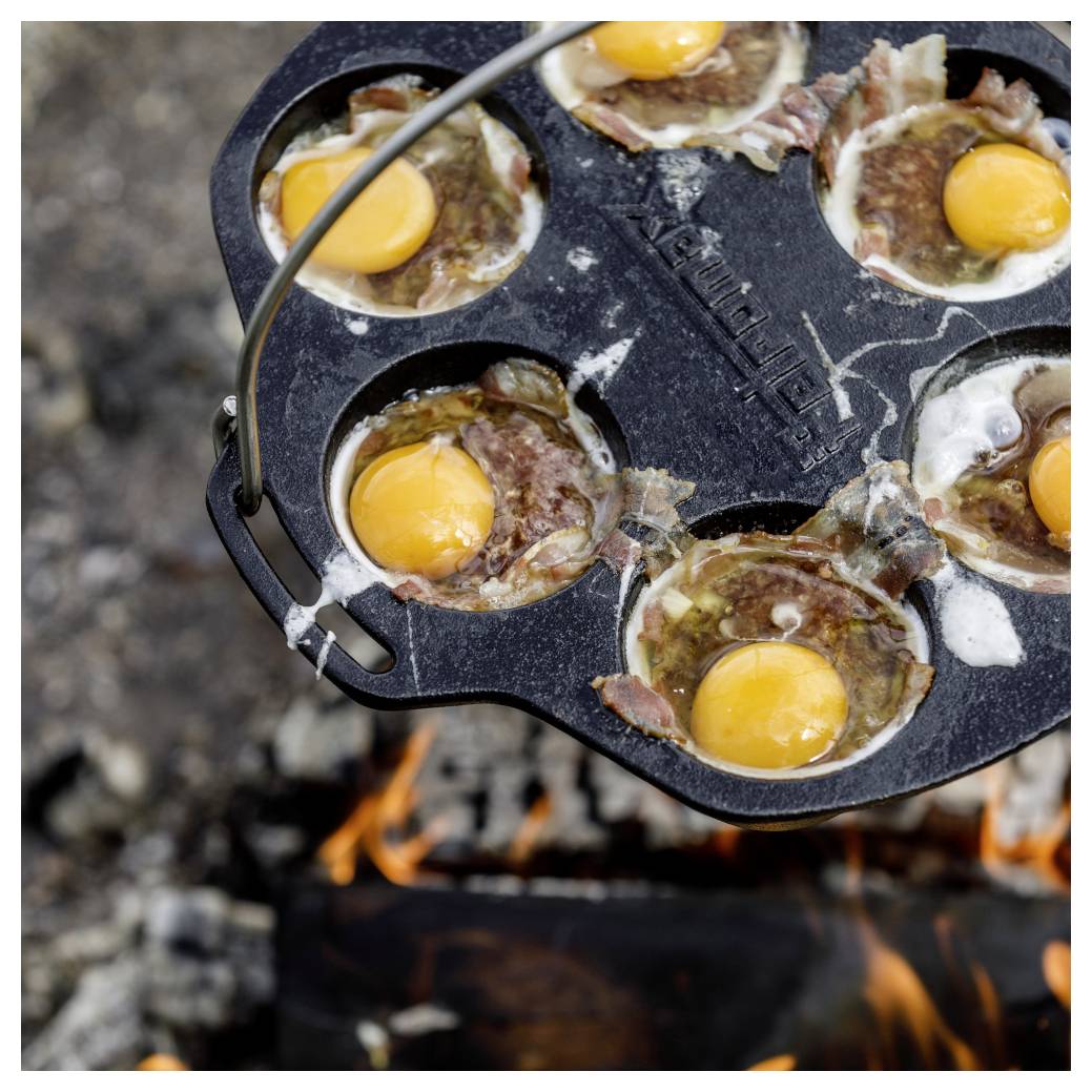 Gusseiserne Pfanne über einem Lagerfeuer mit mehreren aufgeschlagenen Eiern und Speck, die im Freien kochen.