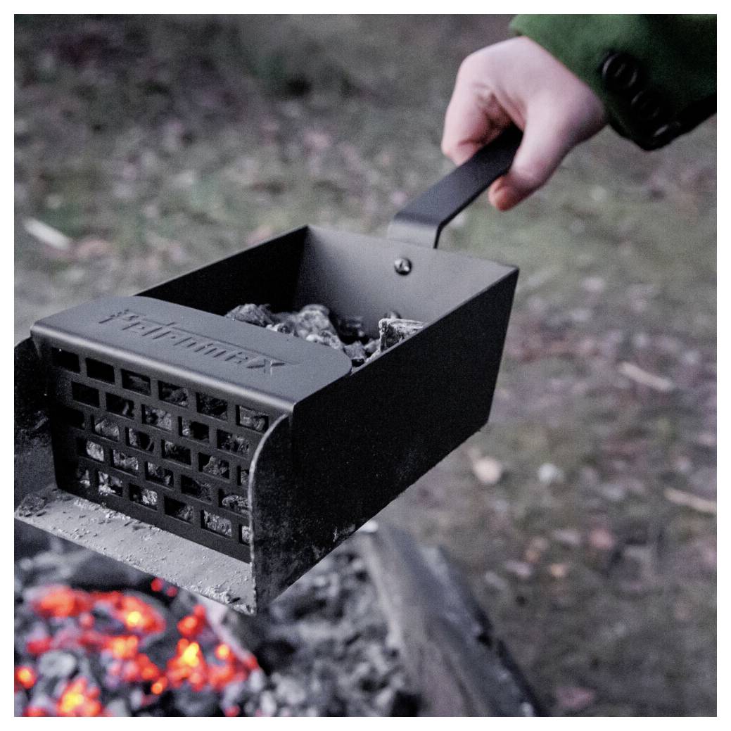 Hand hält einen Metallgrillanzünder gefüllt mit heißen Kohlen über einem Lagerfeuer, bereit, um einen Grill oder eine Feuerstelle anzuzünden.