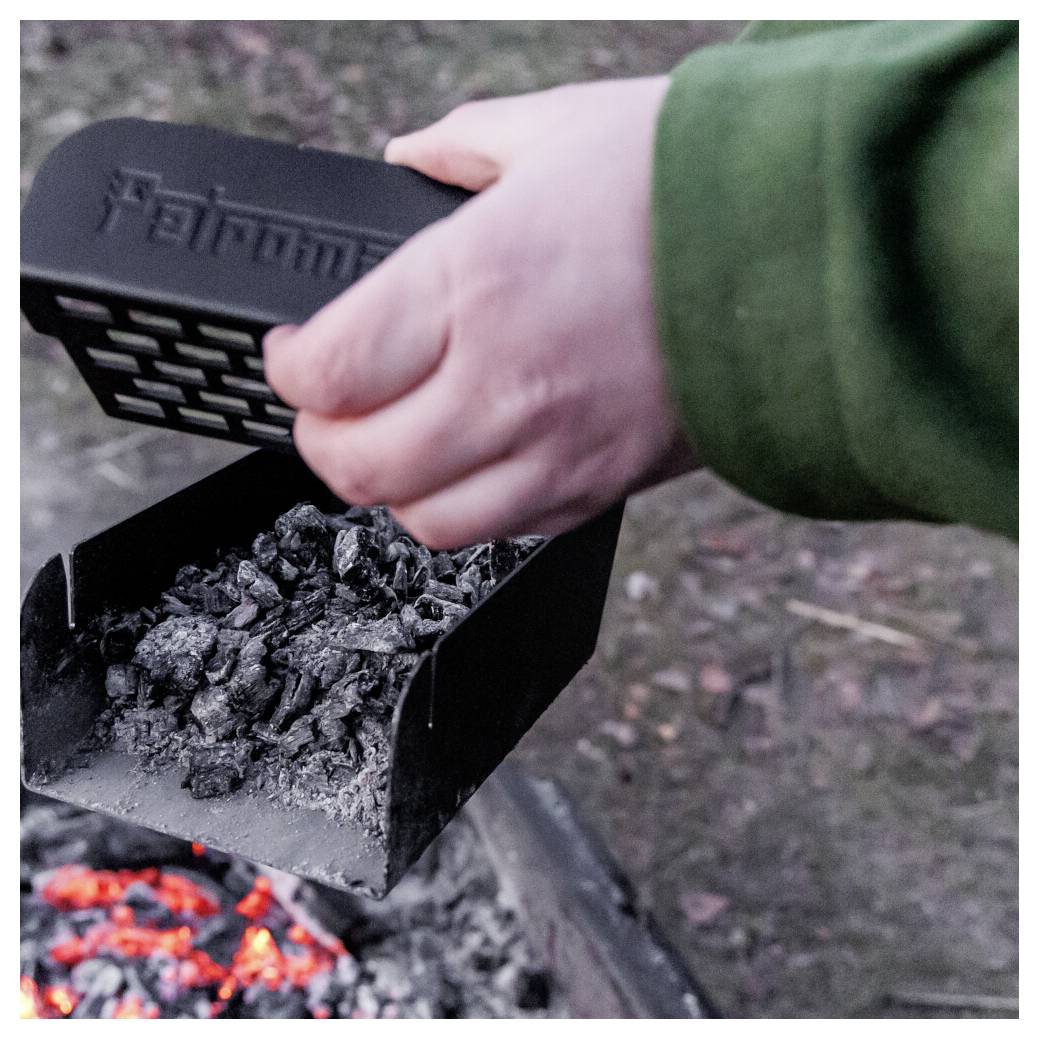 Eine Person gießt Holzkohle aus einem schwarzen Metallkohleanzünder in eine Feuerstelle mit glühenden Gluten darunter.