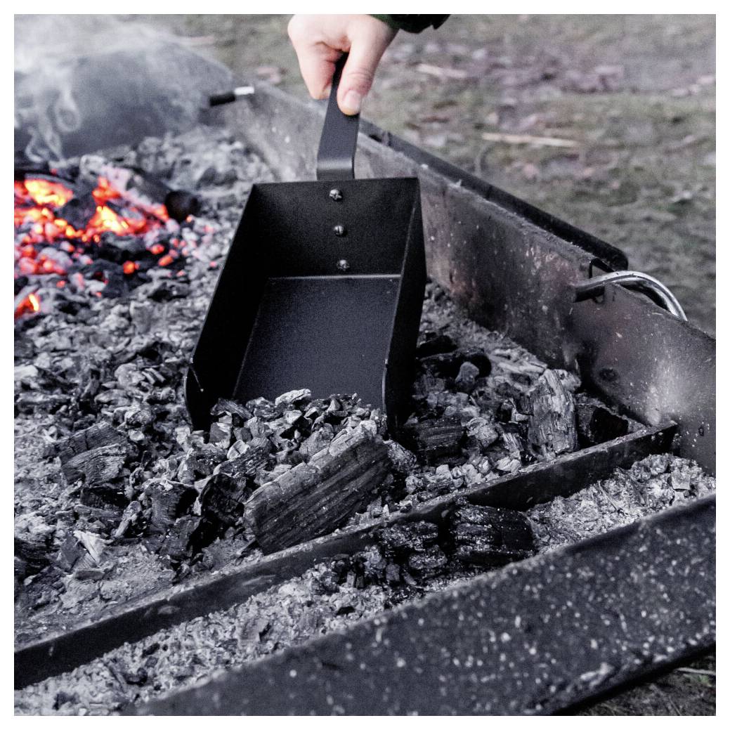 Hand, die einen schwarzen Metallaschenaufschieber verwendet, um glühende Kohlen aus einer Feuerstelle zu schaufeln, mit leuchtenden Gluten, die neben verkohltem Holz sichtbar sind.