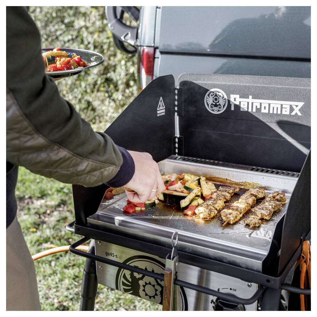 Person kocht Spieße und Gemüse auf einem tragbaren Petromax-Gasgrill im Freien, mit einem Teller mit Essen in der Nähe.
