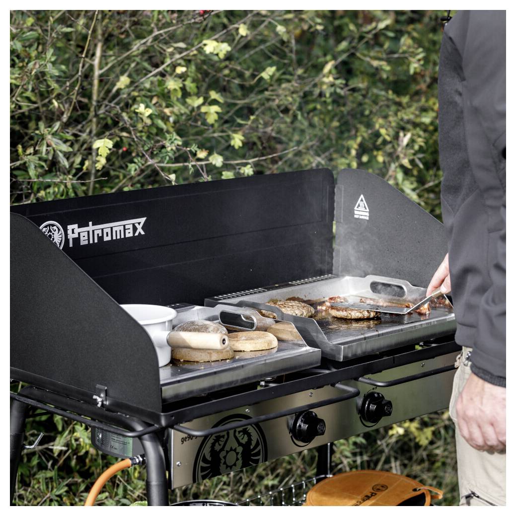 Person, die Burger und Brötchen auf einem Petromax-Gasgrill im Freien grillt, mit einem Spatel und sichtbaren Seitentabletts.