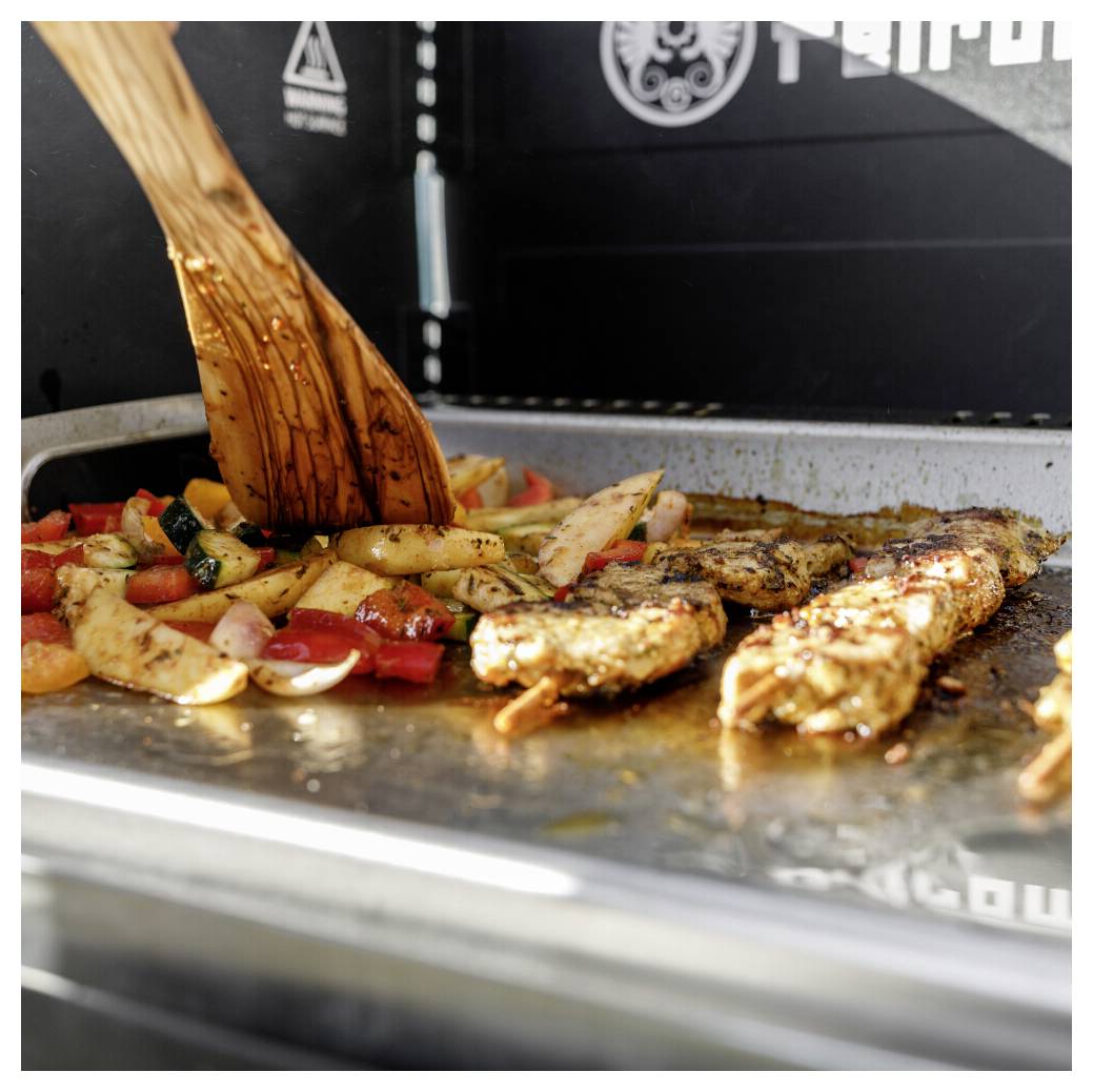 Hühnchenspieße und geschnittenes Gemüse grillen auf einem Flachbratgrill, mit einem Holzspatel, der das Essen wendet.