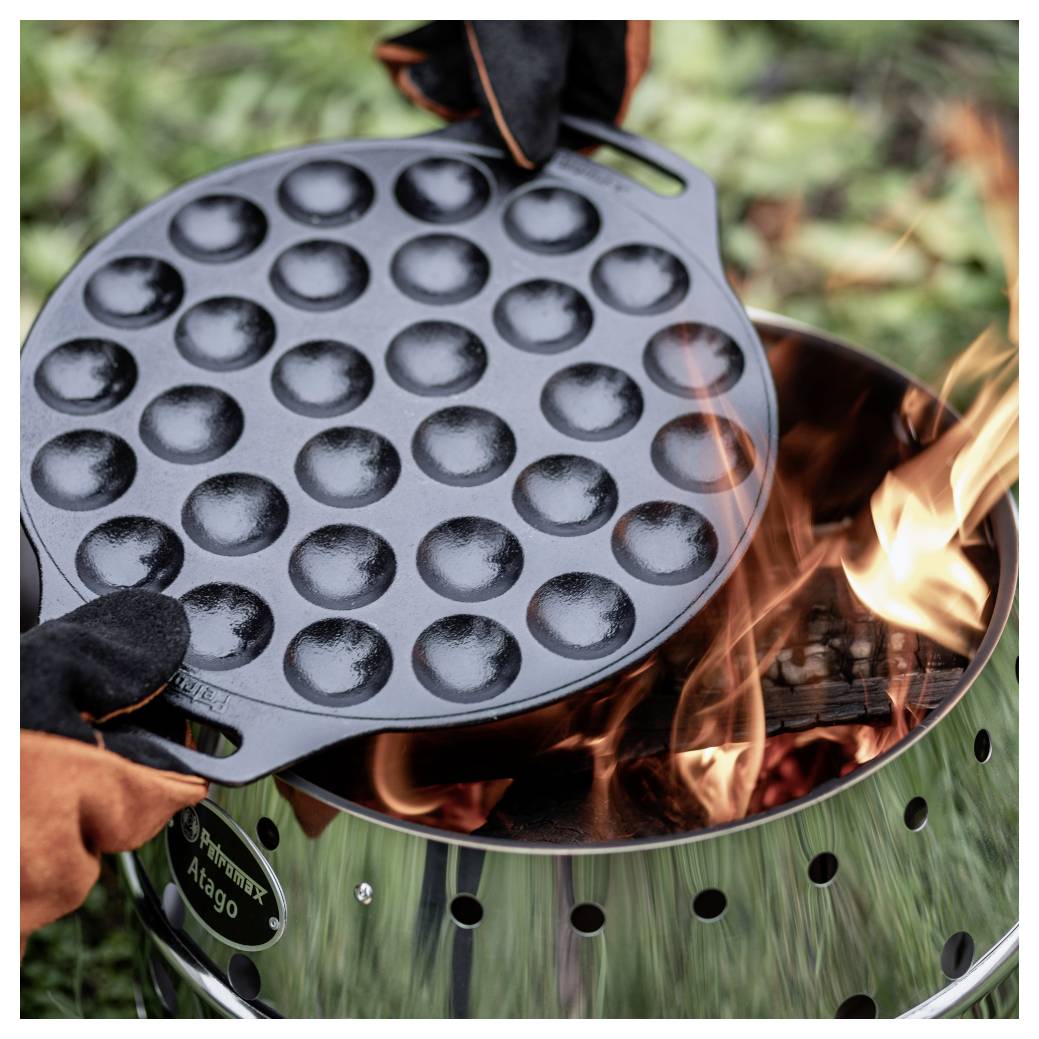 Hände heben eine runde Gusseisenpfanne mit Teigballen von einem brennenden Außengrill oder Lagerfeuer.