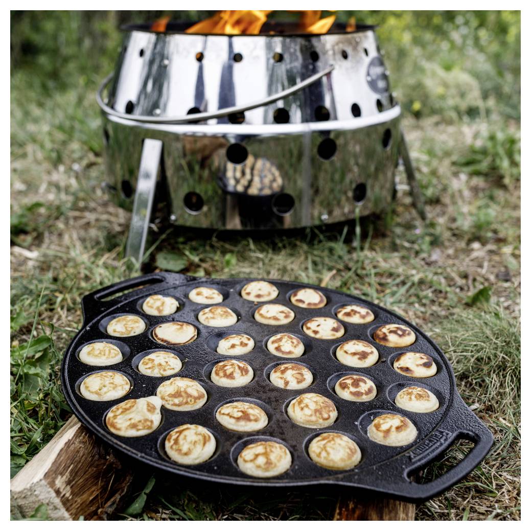 Outdoor-Kochszene mit einer gusseisernen Platte voller kleiner Pfannkuchen oder Blini auf Gras neben einer brennenden Metallfeuer-Feuerstelle.