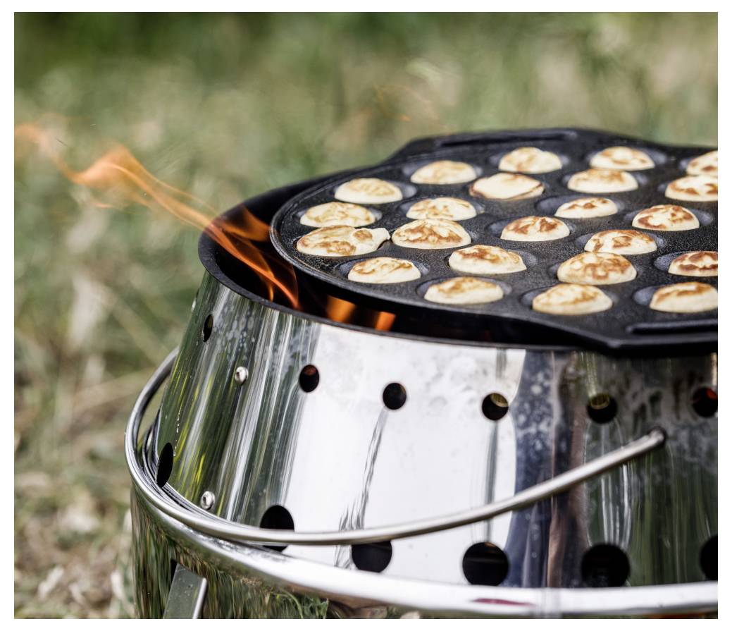 Kleine Pfannkuchen braten auf einer runden Grillplatte über einem Outdoor-Holzkohlengrill mit sichtbaren Flammen.