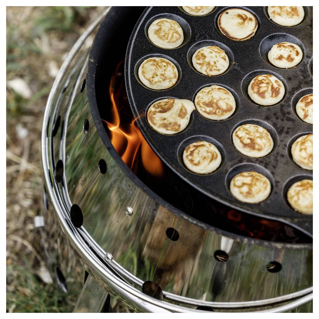 Mini-Pfannkuchen-Teig, der in einer runden Gusseisenpfanne über einer offenen Flamme auf einem Grill gekocht wird.