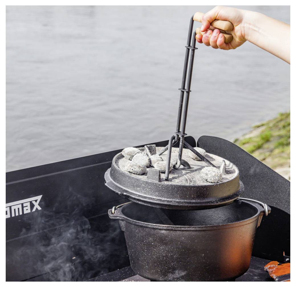 Hand hebt ein Grillwerkzeug, um einen heißen Deckel von einem Räucherofen oder Topf neben Wasser zu entfernen, mit aufsteigendem Dampf.