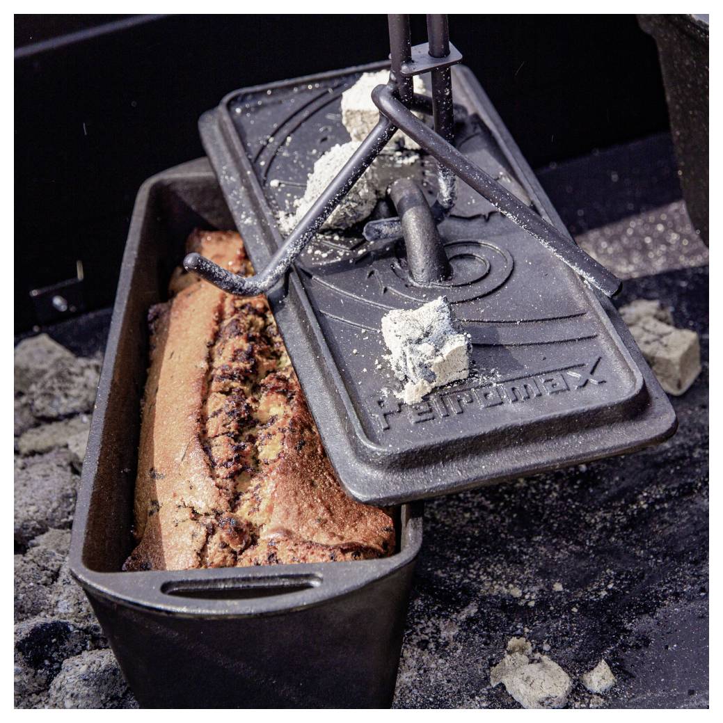 Gusseiserner Dutch Oven über Kohlen mit angehobenem Deckel, der einen im Inneren backenden Brotlaib offenbart.