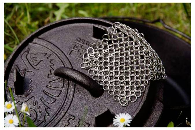 Nahaufnahme eines Deckelgriffs aus Gusseisen oder eines Dutch-Oven-Deckels mit einer Kettenmail-Reinigungsbürste oben drauf, im Freien auf Gras mit kleinen weißen Blüten angeordnet.