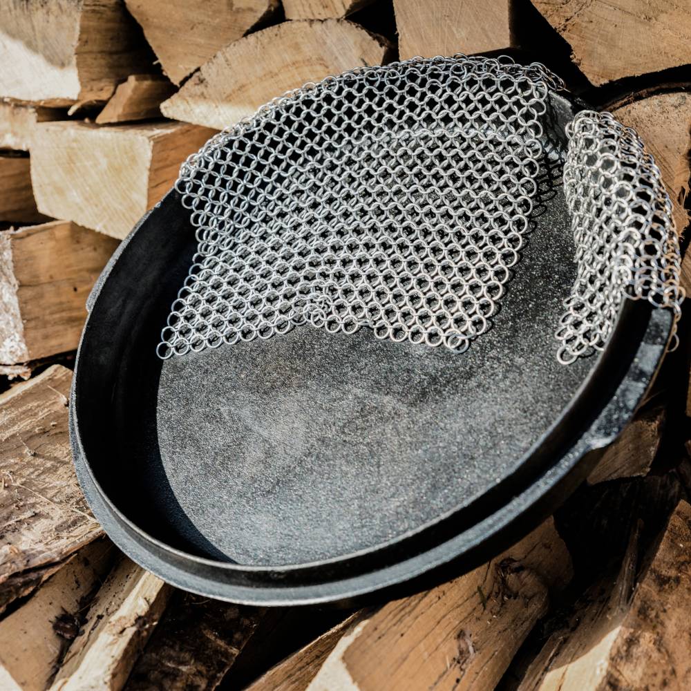 Runde schwarze Pfanne oder Hut mit silbernem kettenhemdähnlichem Netzgewebe darüber drapiert, ruhend auf gestapeltem Brennholz.