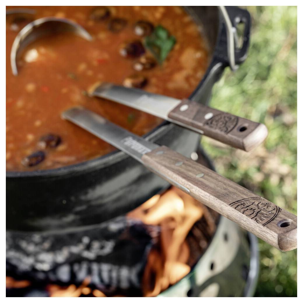 Zwei Messer mit Holzgriffen ruhen auf einem Topf mit köchelndem Chili auf einem Campingkocher über offenem Feuer im Freien.