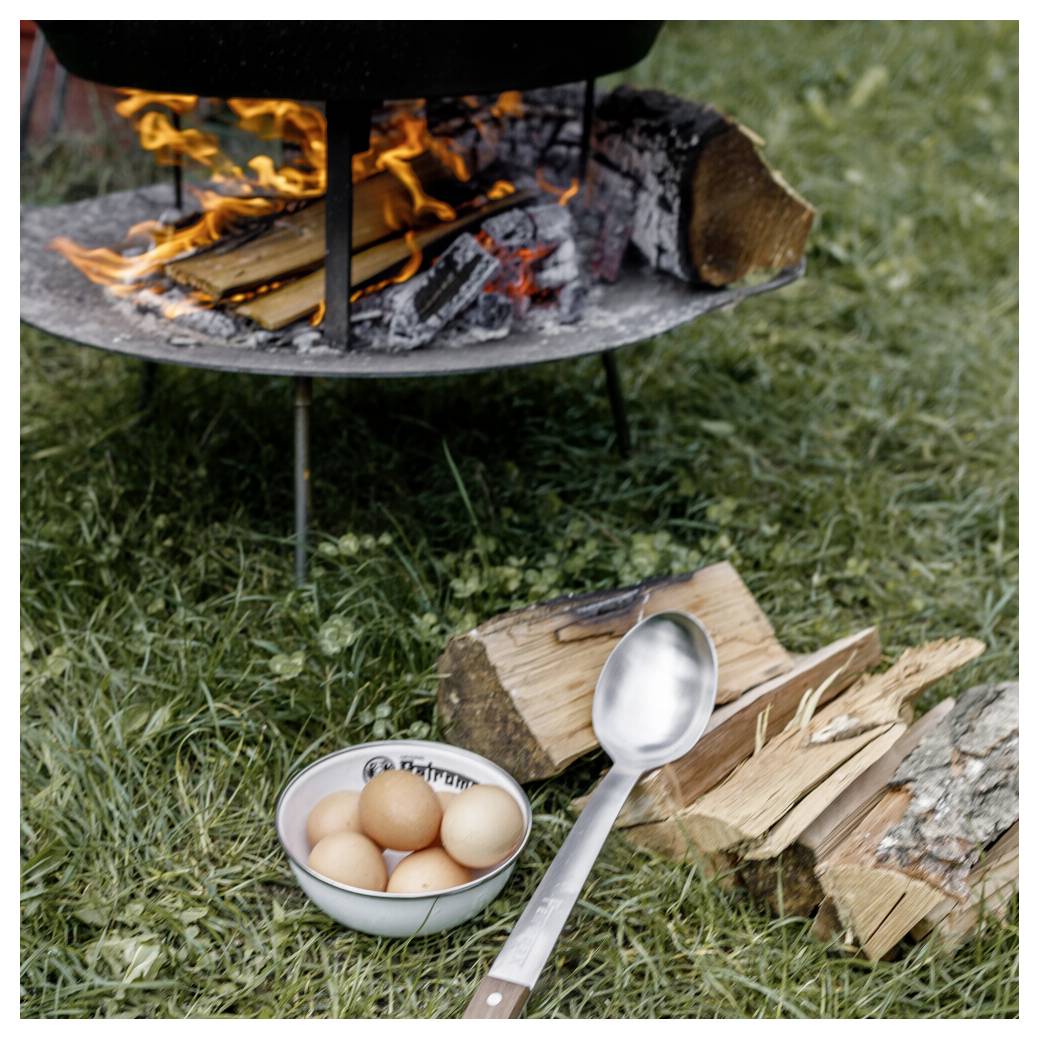Ein Lagerfeuer brennt in einer Metallfeuergrube neben gespaltenem Feuerholz, einer Schüssel mit Eiern und einem Löffel auf Gras im Freien.
