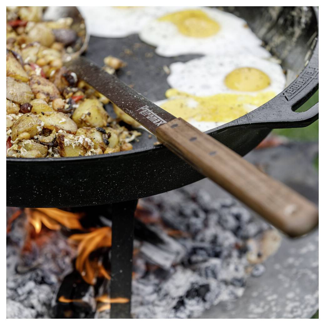 Spiegeleier und Kartoffeln, die in einer großen Pfanne über einem offenen Lagerfeuer kochen, mit einem Spachtel, der über die Pfanne gelegt ist.