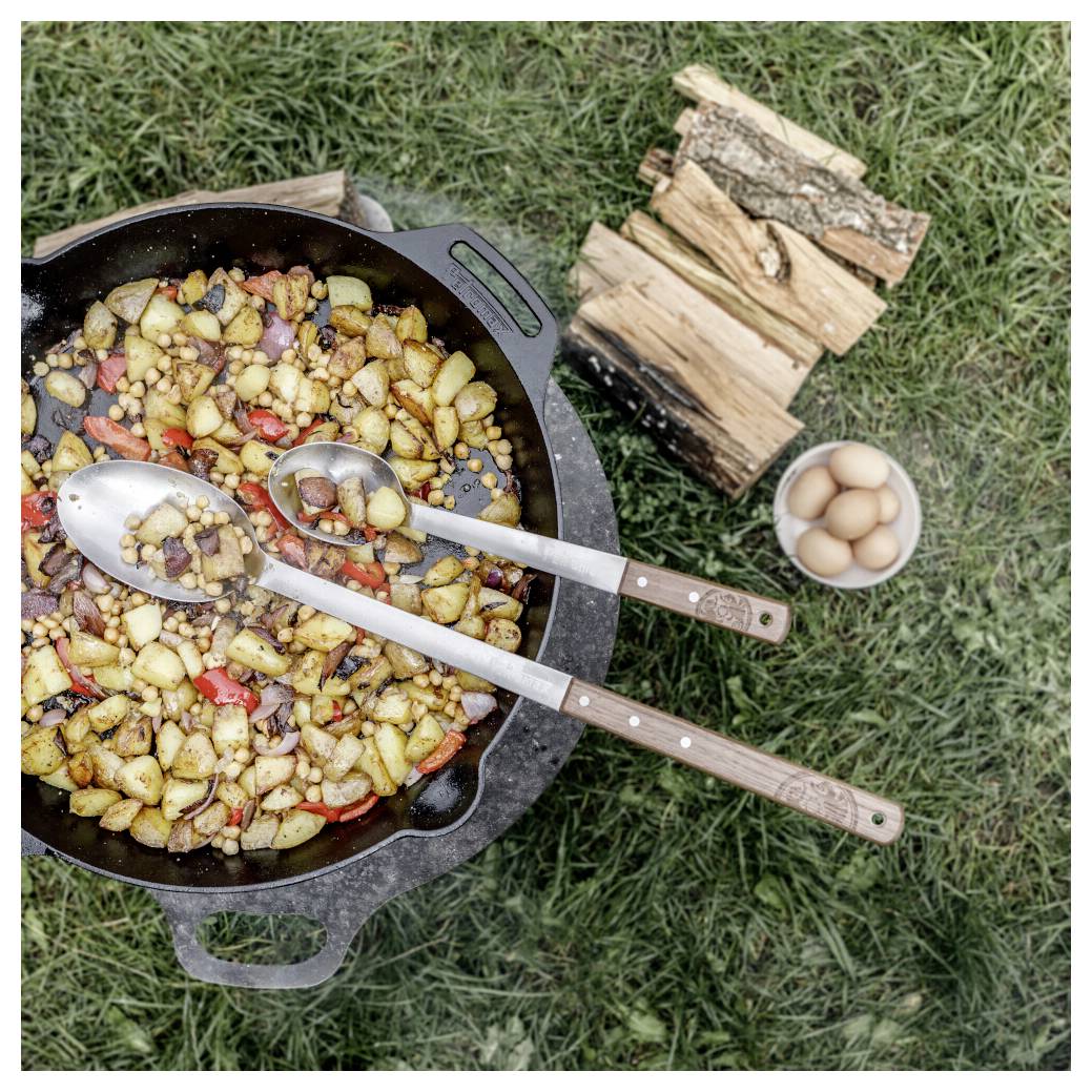 Gusseiserne Pfanne mit sautiertem Kartoffeln und Gemüse, zwei Holzlöffel, gestapeltes Brennholz und eine Schüssel mit Eiern auf Gras.