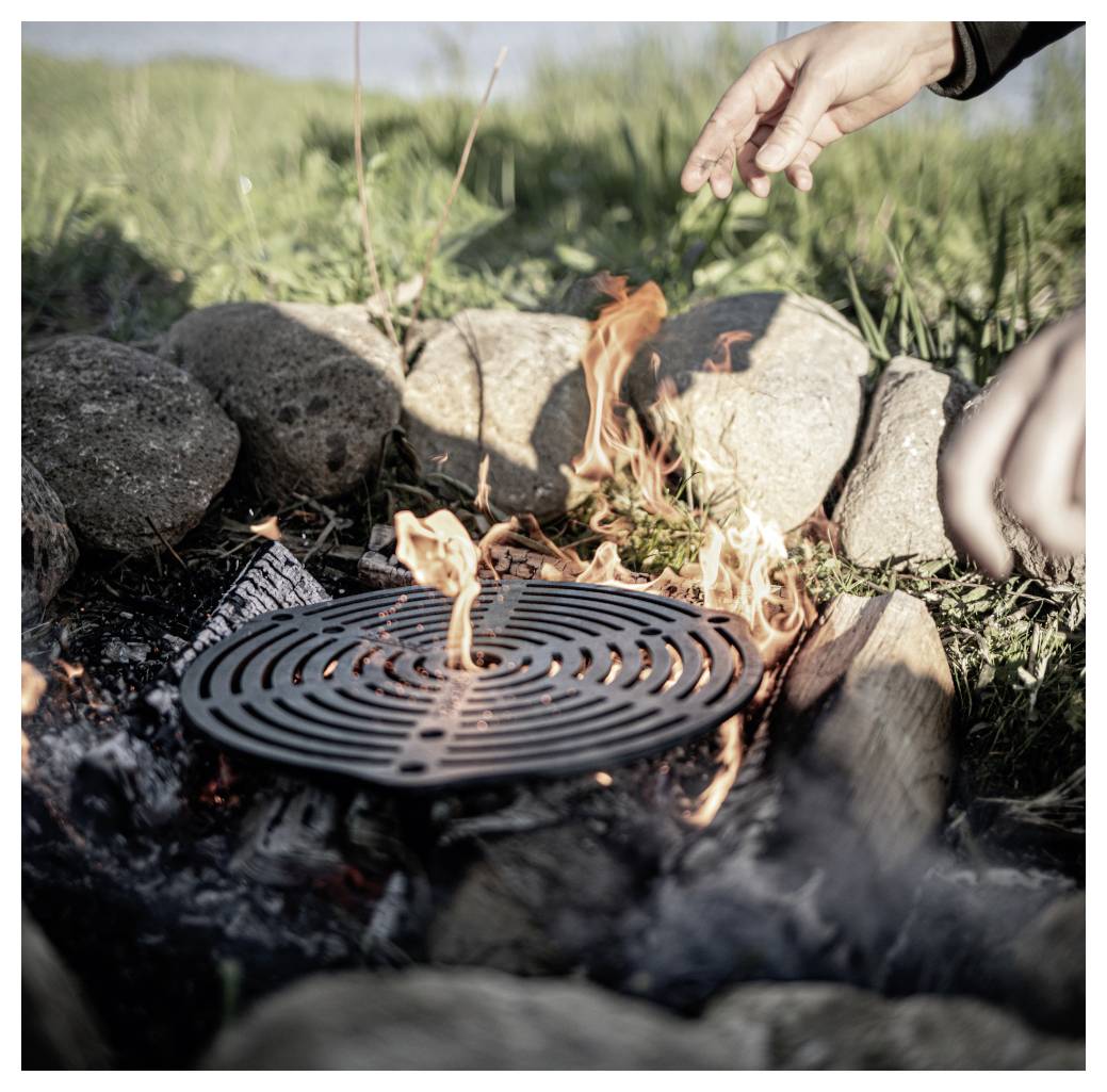 Eine Hand, die sich einem kleinen Lagerfeuer nähert, das unter einem runden Metallrost brennt, umgeben von Steinen im Freien.