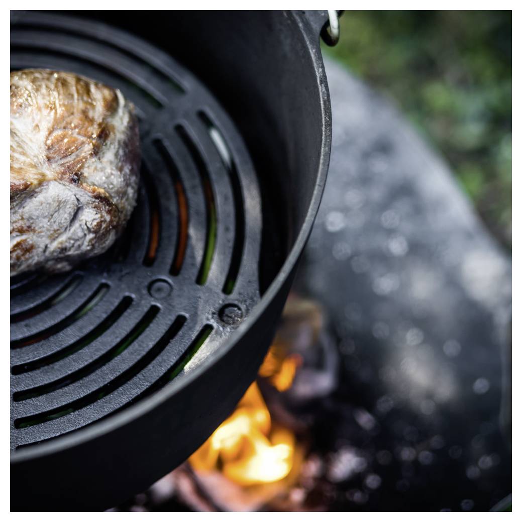 Nahaufnahme eines gusseisernen Topfes über offenem Feuer, mit einem angebratenen Stück Fleisch oder Brot, das darin kocht.