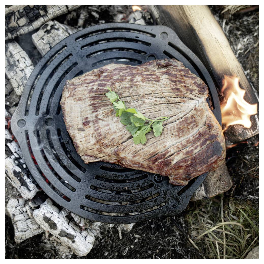 Gegrilltes Steak, das auf einem runden Gusseisenrost über einem offenen Lagerfeuer angebraten wird, belegt mit einem kleinen Kraut- oder Kräuterzweig.