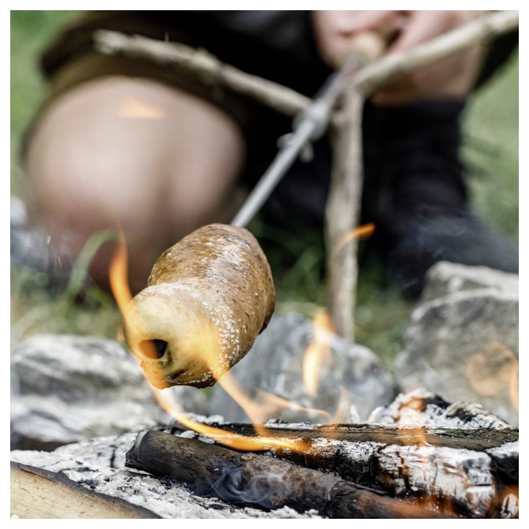 Person, die Brotteig auf einem Stock über einem offenen Lagerfeuer röstet, mit sichtbaren Flammen und Glut.