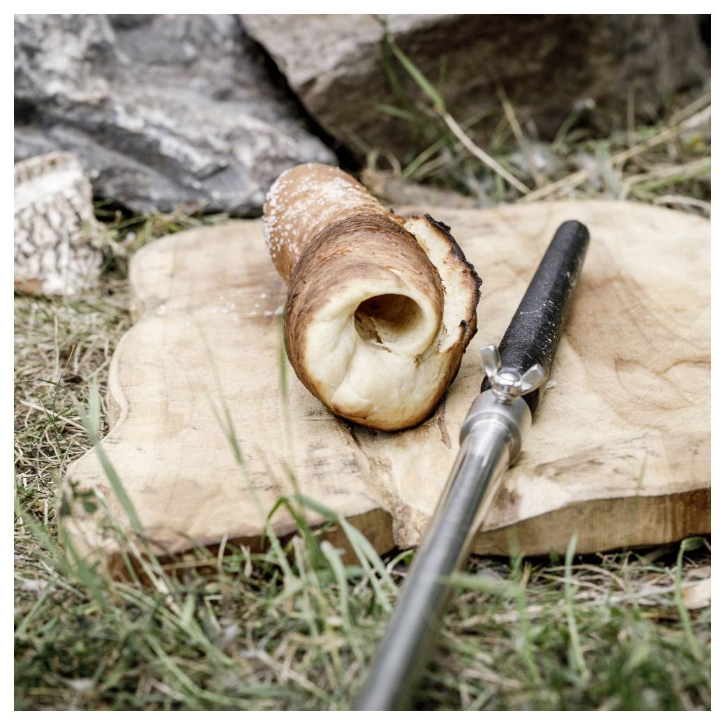 Ein gerösteter Brotkegel auf einem Holzbrett neben einem Messer, auf Gras im Freien angeordnet.