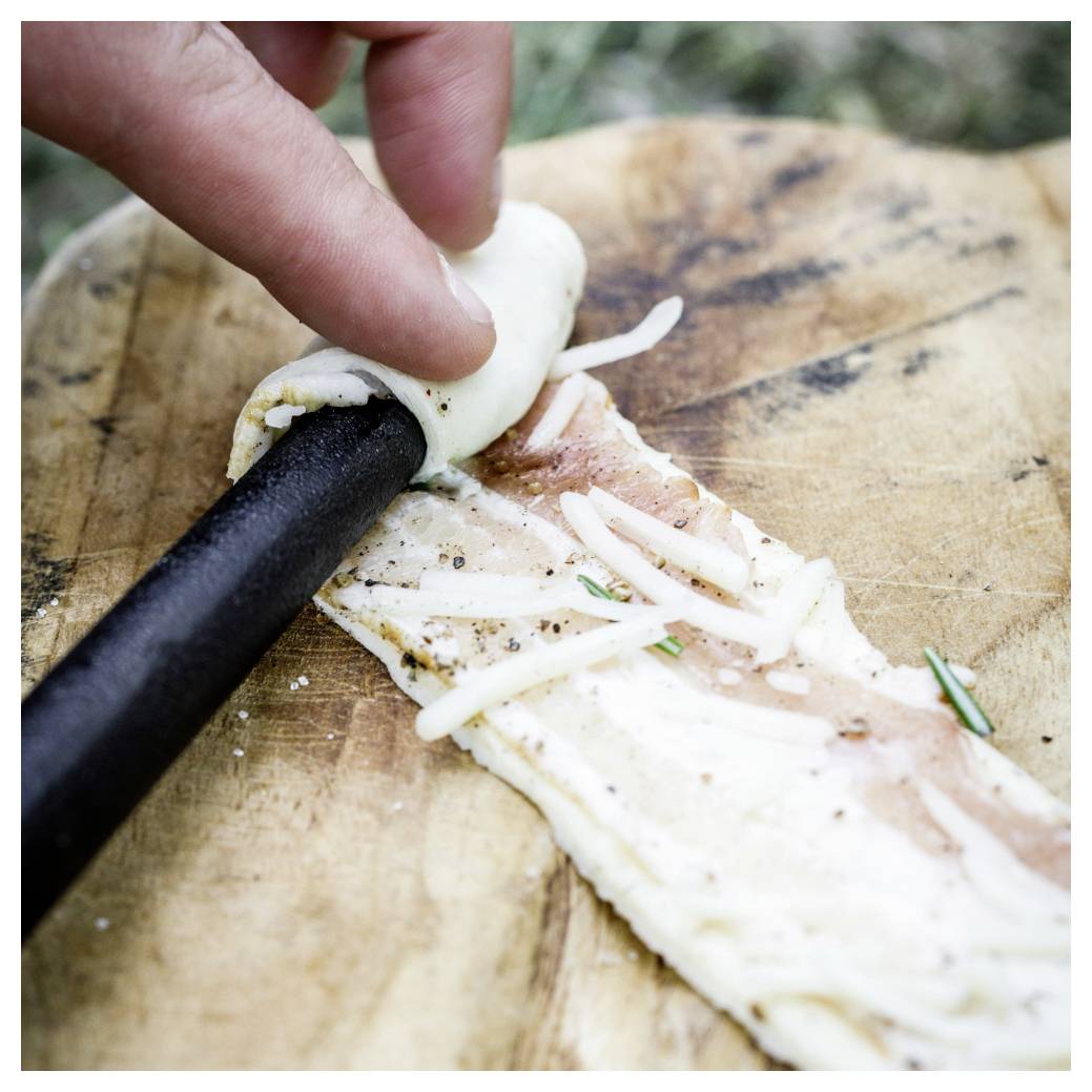 Eine dünne Scheibe rohen Fisches wird von Hand um einen schwarzen Stab mit feingeschnittenen Gemüse auf einem Holzschneidebrett gerollt.