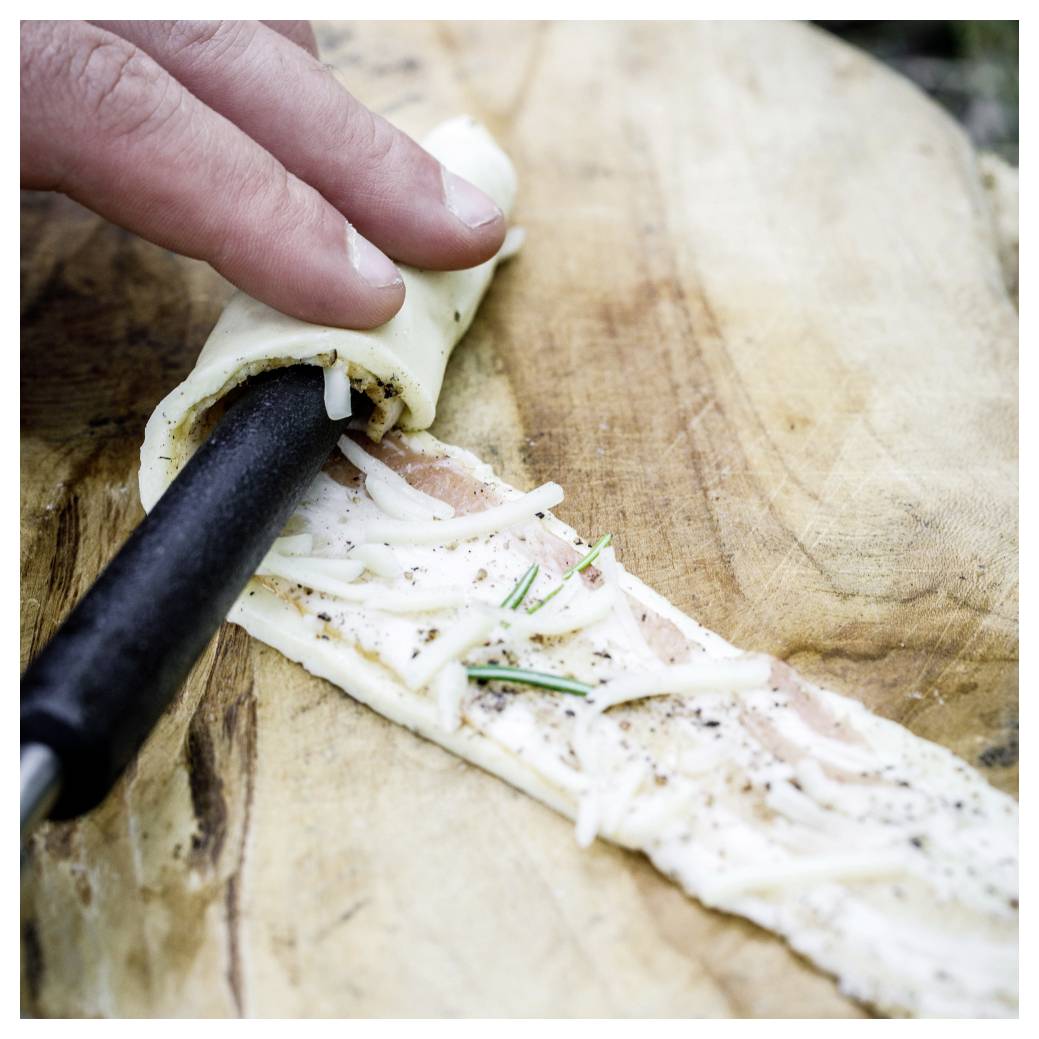 Die Hand wickelt ein Fladenbrot mit Käse und Kräutern um ein schwarzes Kochgerät auf einem Holzbrett.