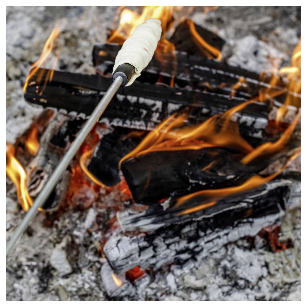 Ein Spieß mit weißem Teig wird über einem offenen Lagerfeuer geröstet, mit Flammen und verkohlten Holzstücken, die sichtbar sind.
