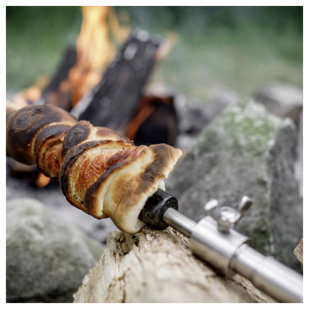 Brotteig auf einem Metallspieß über einem Lagerfeuer gegart, in Spiralen gewickelt und gebräunt, mit Steinen und Flammen im Hintergrund.