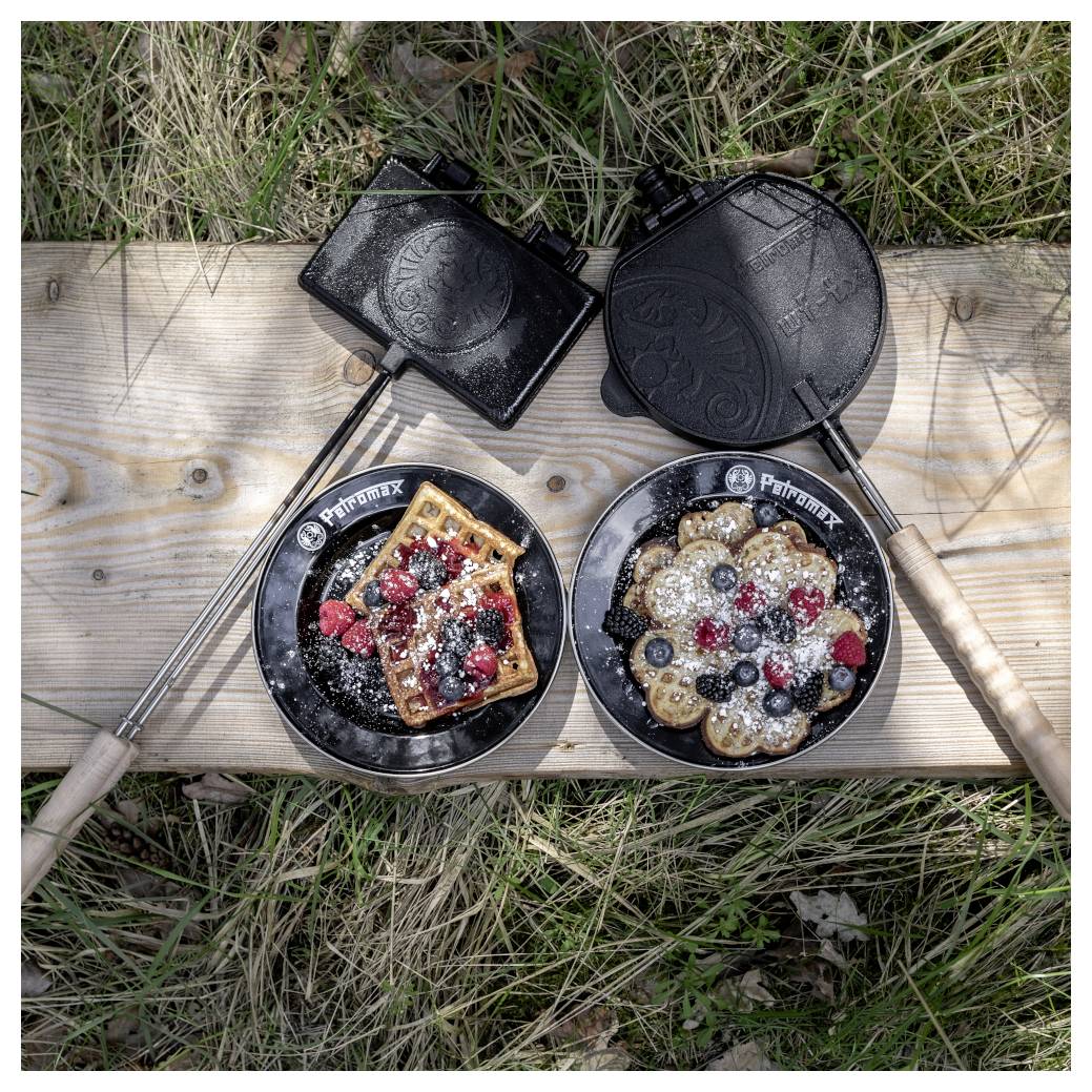 Zwei offene gusseiserne Waffeleisen auf einer hölzernen Planke im Freien, jedes mit einer mit Beeren belegten Waffel, die mit Puderzucker bestäubt ist.