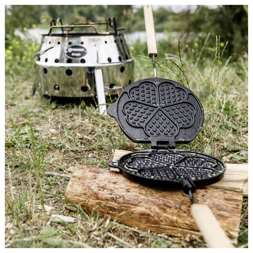 Offene gusseiserne Waffeleisen auf einem Baumstamm über Gras, mit einem metallischen Campingkocher dahinter.