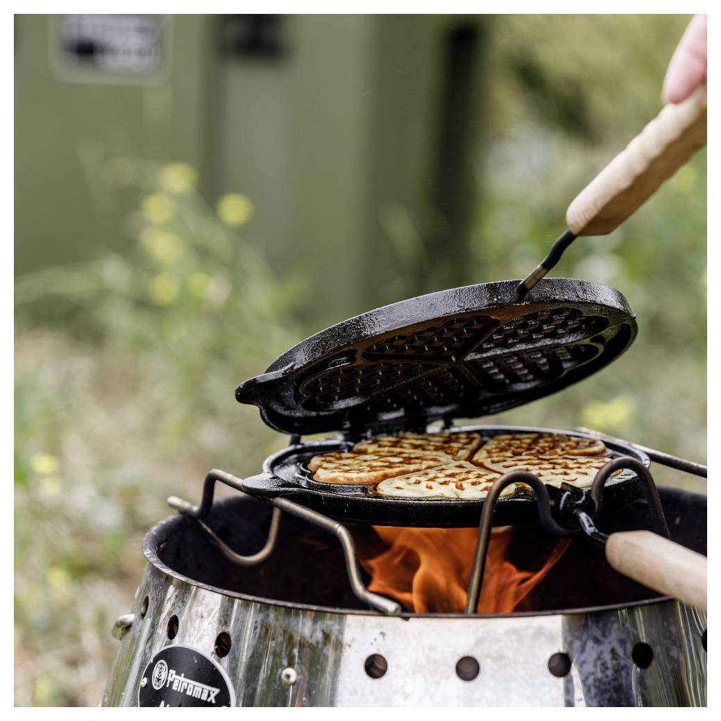 Eine Person benutzt ein Waffeleisen über einem Lagerfeuer im Freien und backt Waffeln über sichtbaren Flammen.