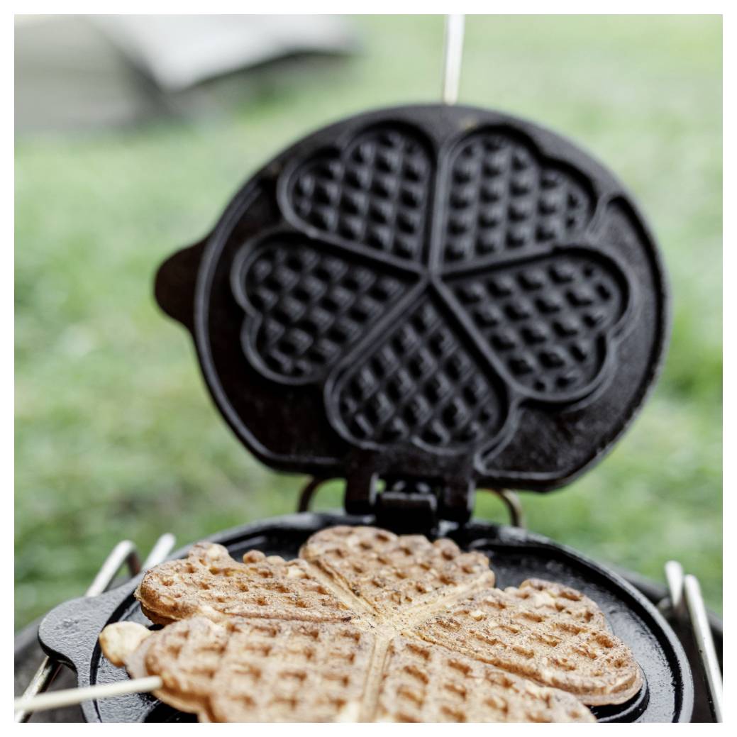 Herzförmiges Waffeleisen, das mehrere goldbraune Waffeln auf einem Küchenherd oder Außengrill zubereitet.