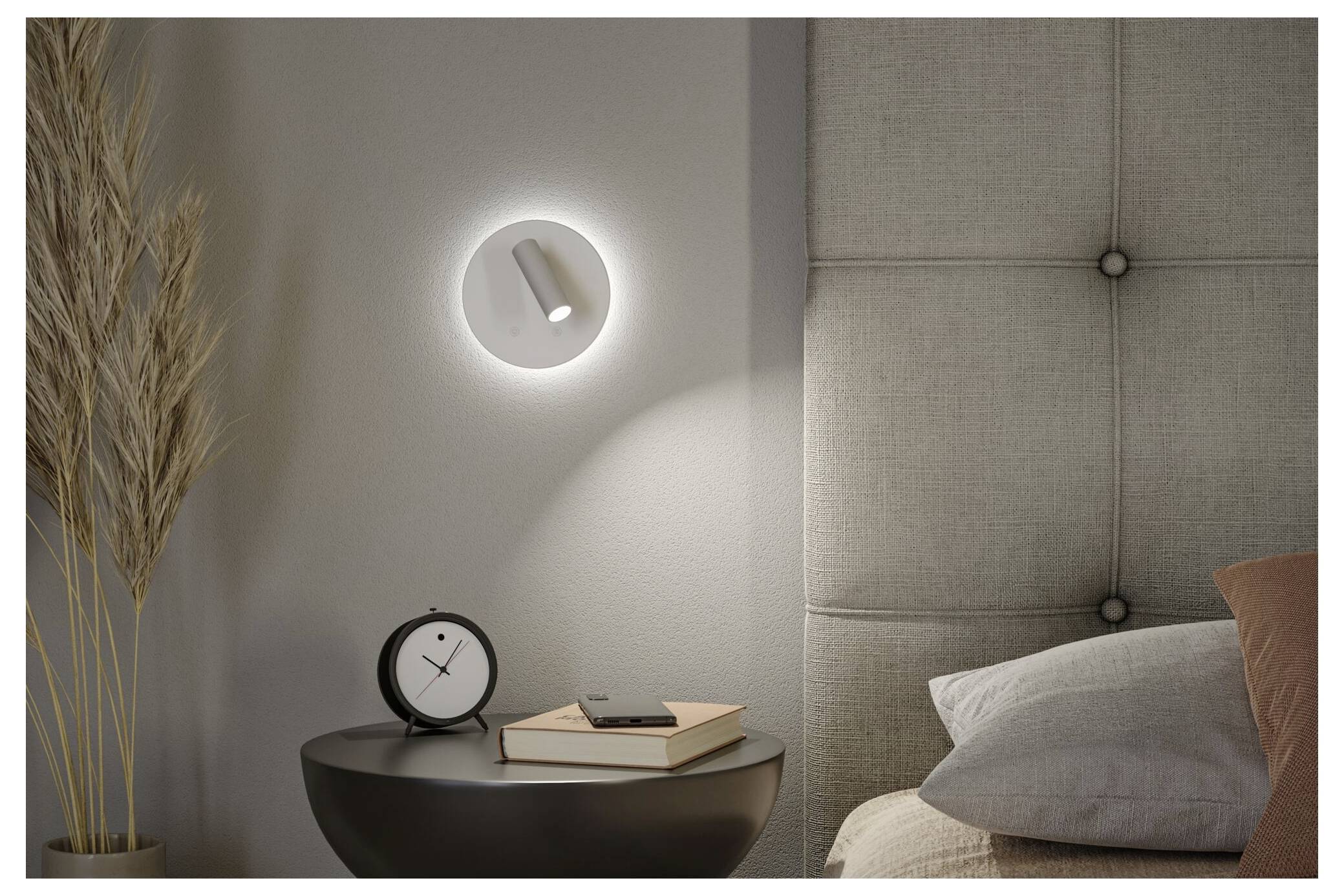 Moderne Schlafzimmerecke mit Wandleuchte, Nachttisch, Uhr, Büchern, Kissen und gepolstertem Kopfteil, warm in der Nacht beleuchtet.