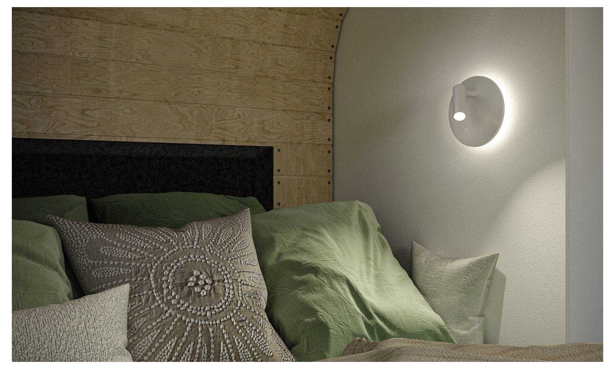 Gemütliches Bett mit grünen und beigen Kissen gegen eine holzgetäfelte Wand, beleuchtet von einer kleinen Wandleuchte.
