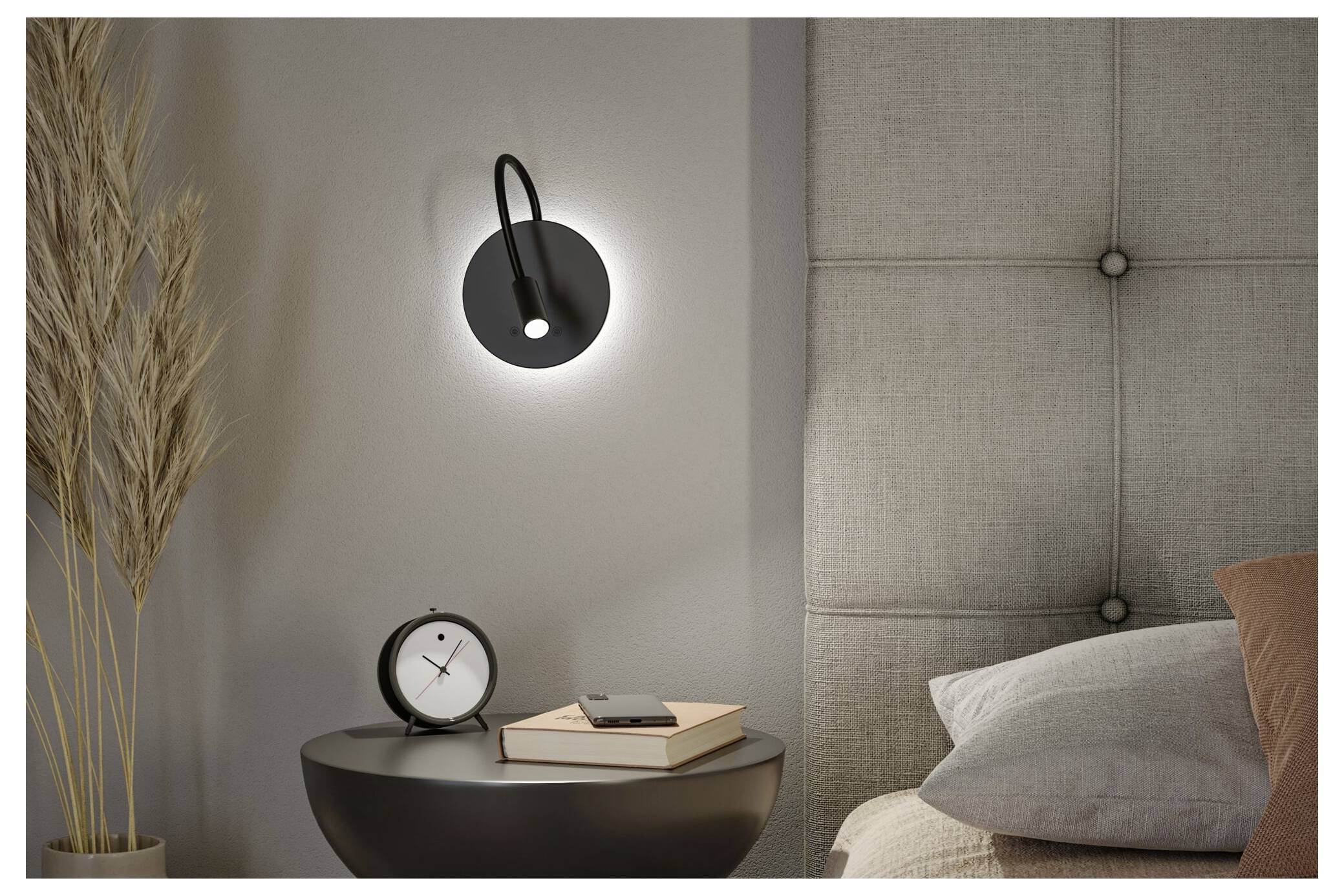 Schlafzimmer-Nachttischszene mit einer an der Wand montierten schwarzen Lampe, kleinem rundem Tisch, Büchern, Uhr, getrockneten Gräsern und Kissen vor einem gesteppten Kopfteil.