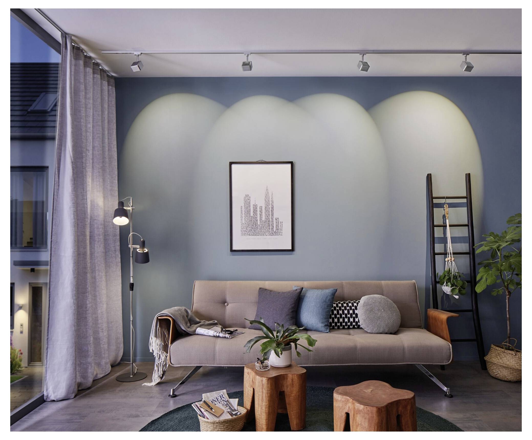 Modernes Wohnzimmer mit beigem Sofa, Stehleuchte, gerahmten Wandbildern, Leiterregalen und Pflanzen vor einer blauen Akzentwand.