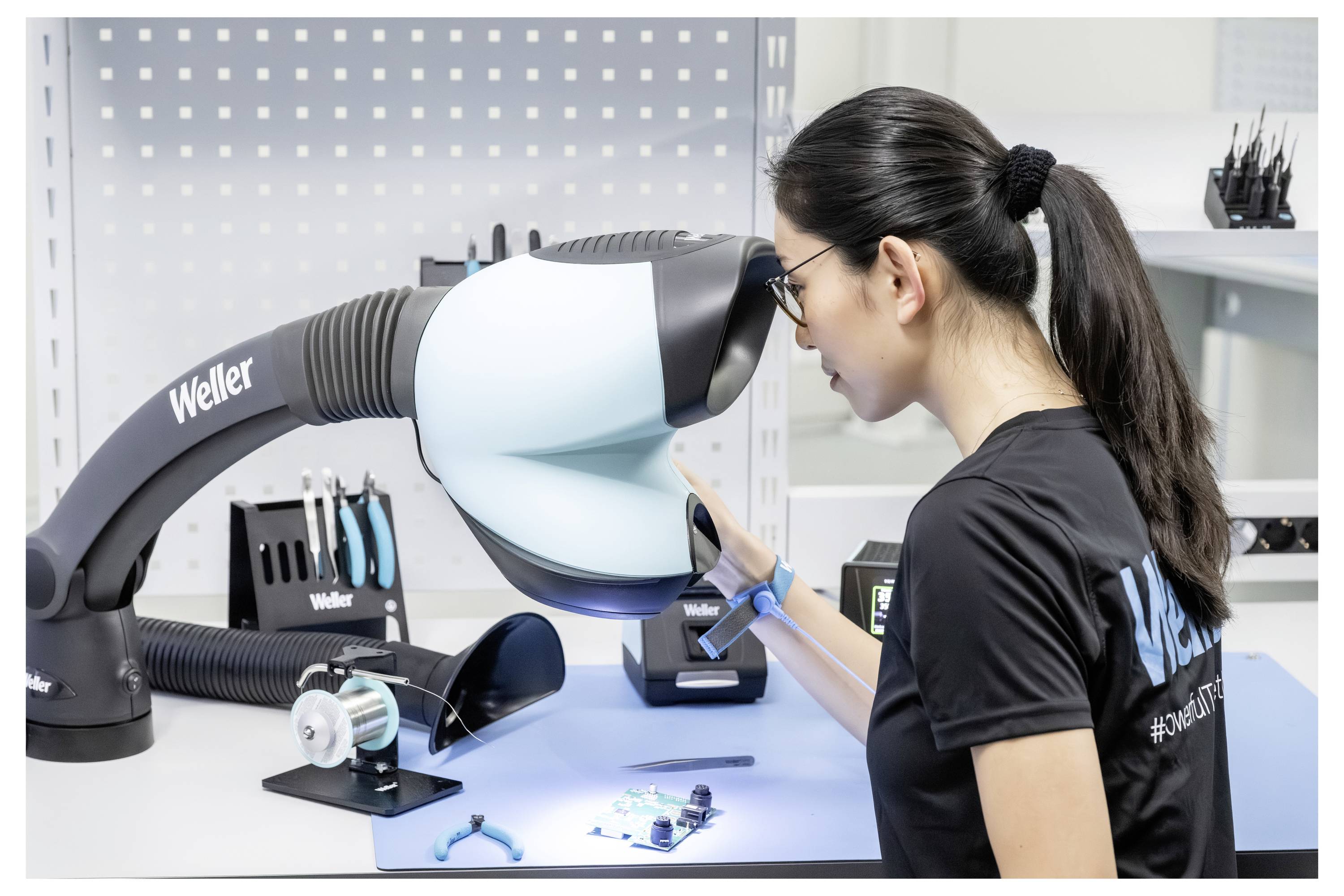 Frau in einem schwarzen Hemd inspiziert ein robotisches Werkbankgerät mit der Bezeichnung Weller in einem sauberen Elektroniklabor.