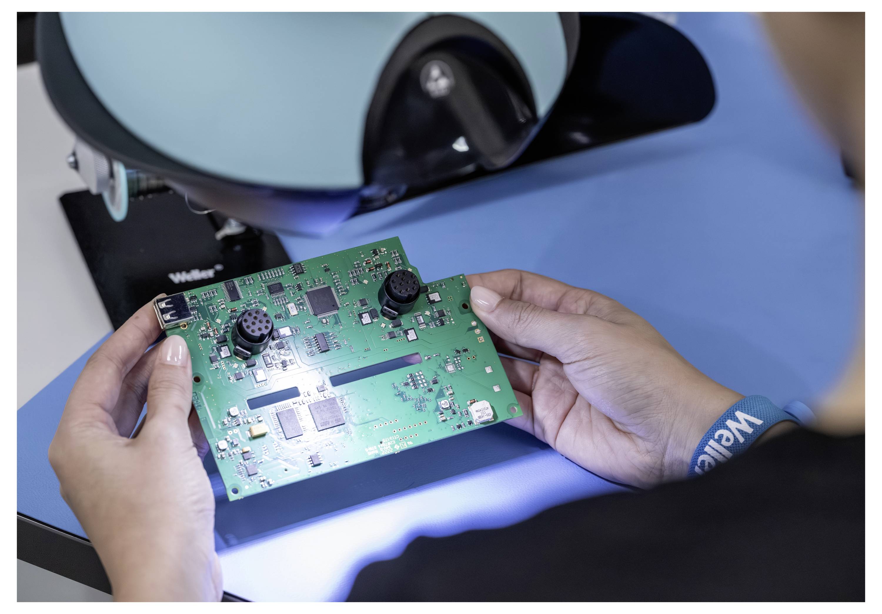 Hände halten eine grüne Leiterplatte unter einem Inspektionsmikroskop, was auf Elektronikqualitätskontrolle oder Reparatur hindeutet.