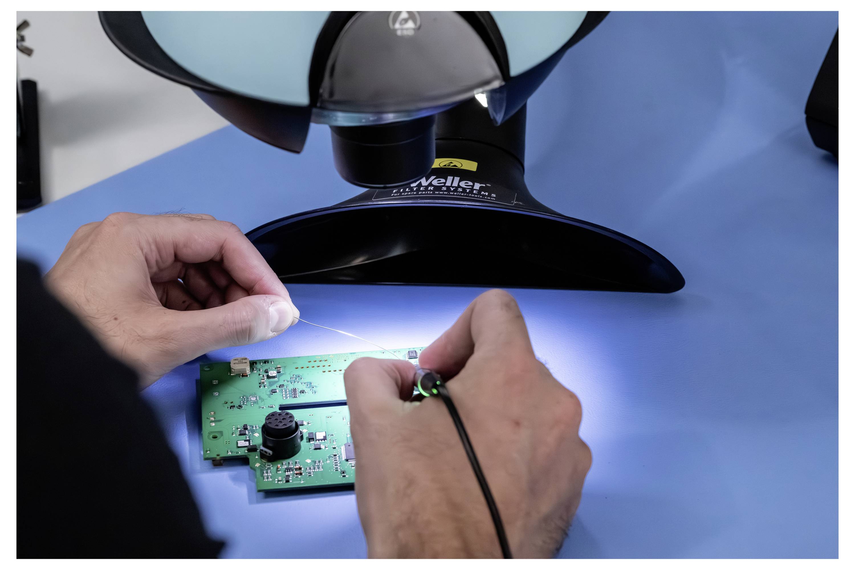 Hände löten eine kleine grüne Leiterplatte unter einem Mikroskop auf einer blauen Arbeitsfläche, was auf Elektronikreparatur oder -inspektion hindeutet.