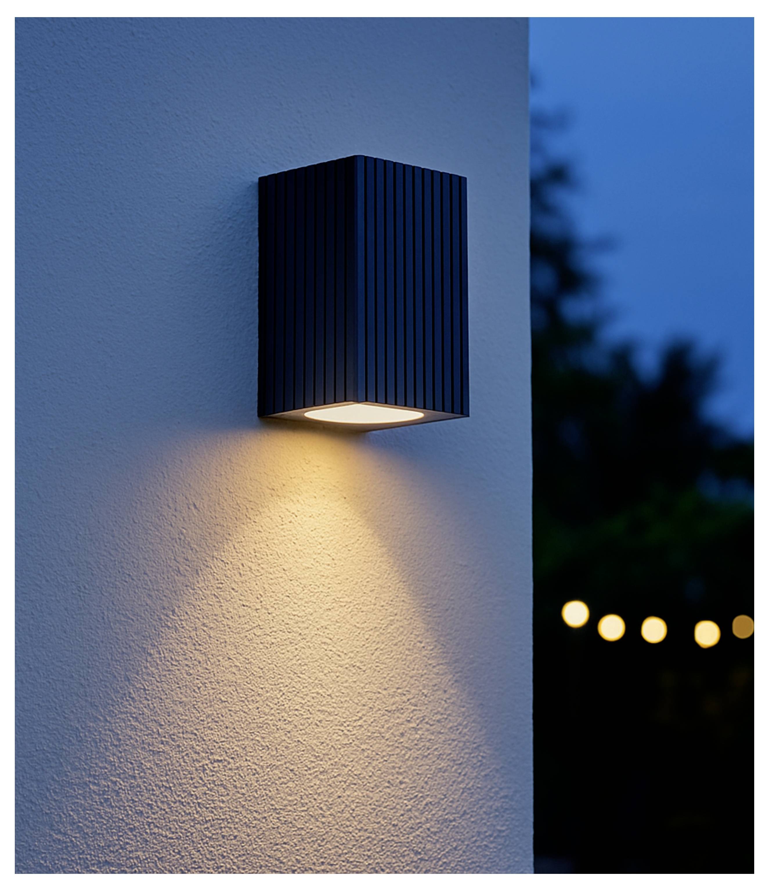 Schwarze Außenwandleuchte wirft einen warmen Lichtkegel auf eine strukturierte Stuckwand in der Dämmerung, mit unscharfen Gartenleuchten im Hintergrund.