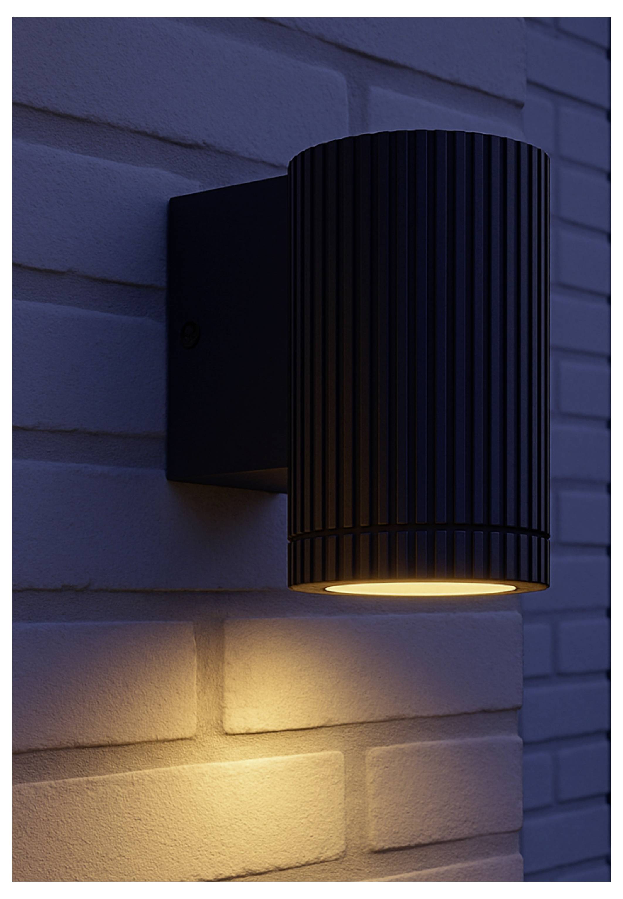 Schwarzer zylindrischer Wandleuchter mit warmem Licht, das nachts eine weiße Backsteinwand beleuchtet.